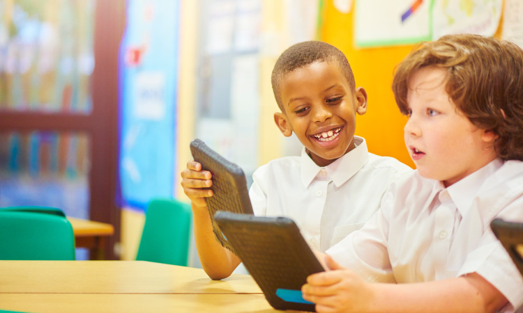 two primary aged schoolchildren sit at their desks and look engrossed in the work that they are doing on their digital tablets. The little boy has an amazed expression whilst looking at his tablet and having fun . They are all wearing white school shirts